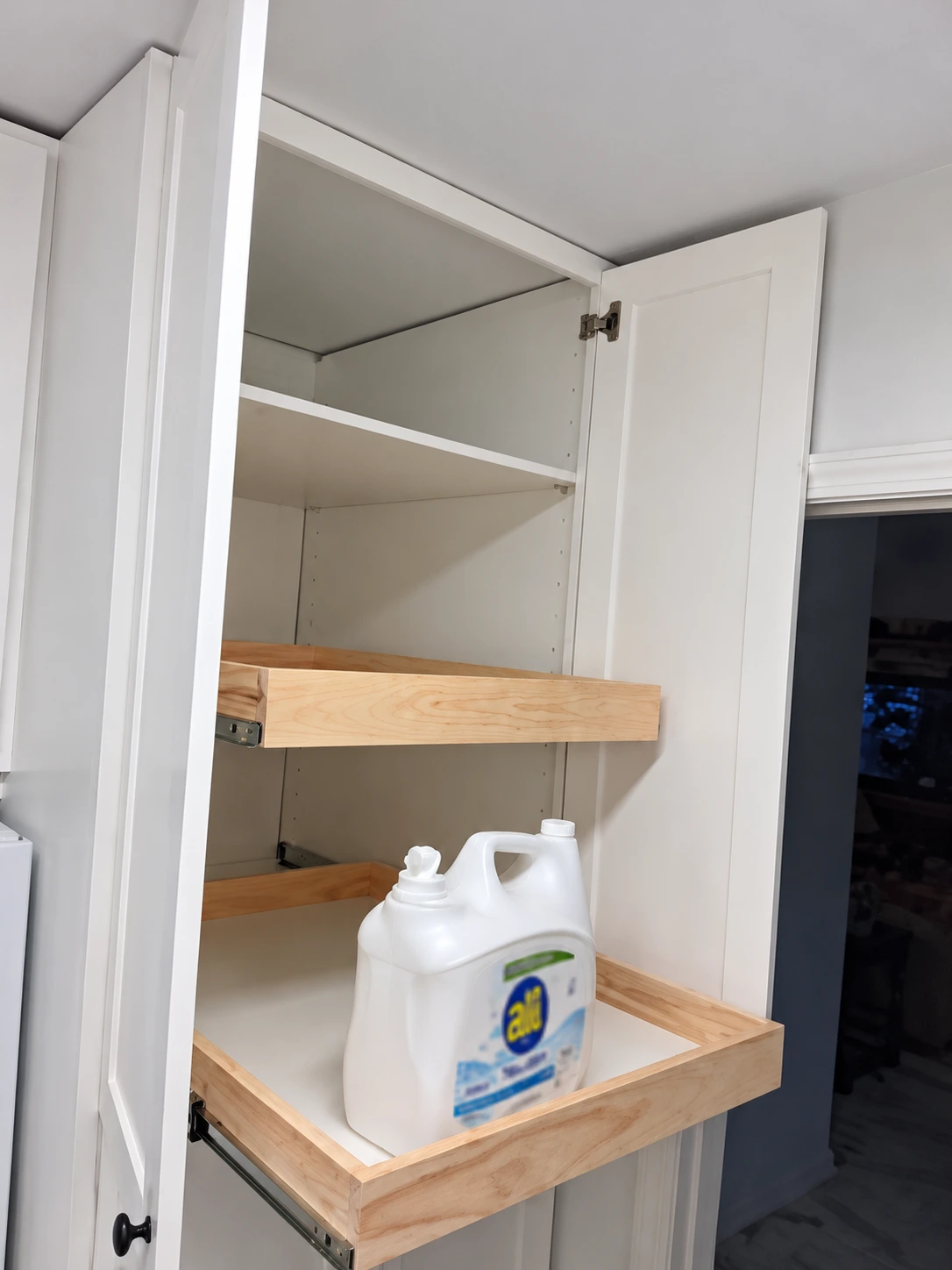 Open white cabinet with wooden pull-out shelves holding cleaning supply container
